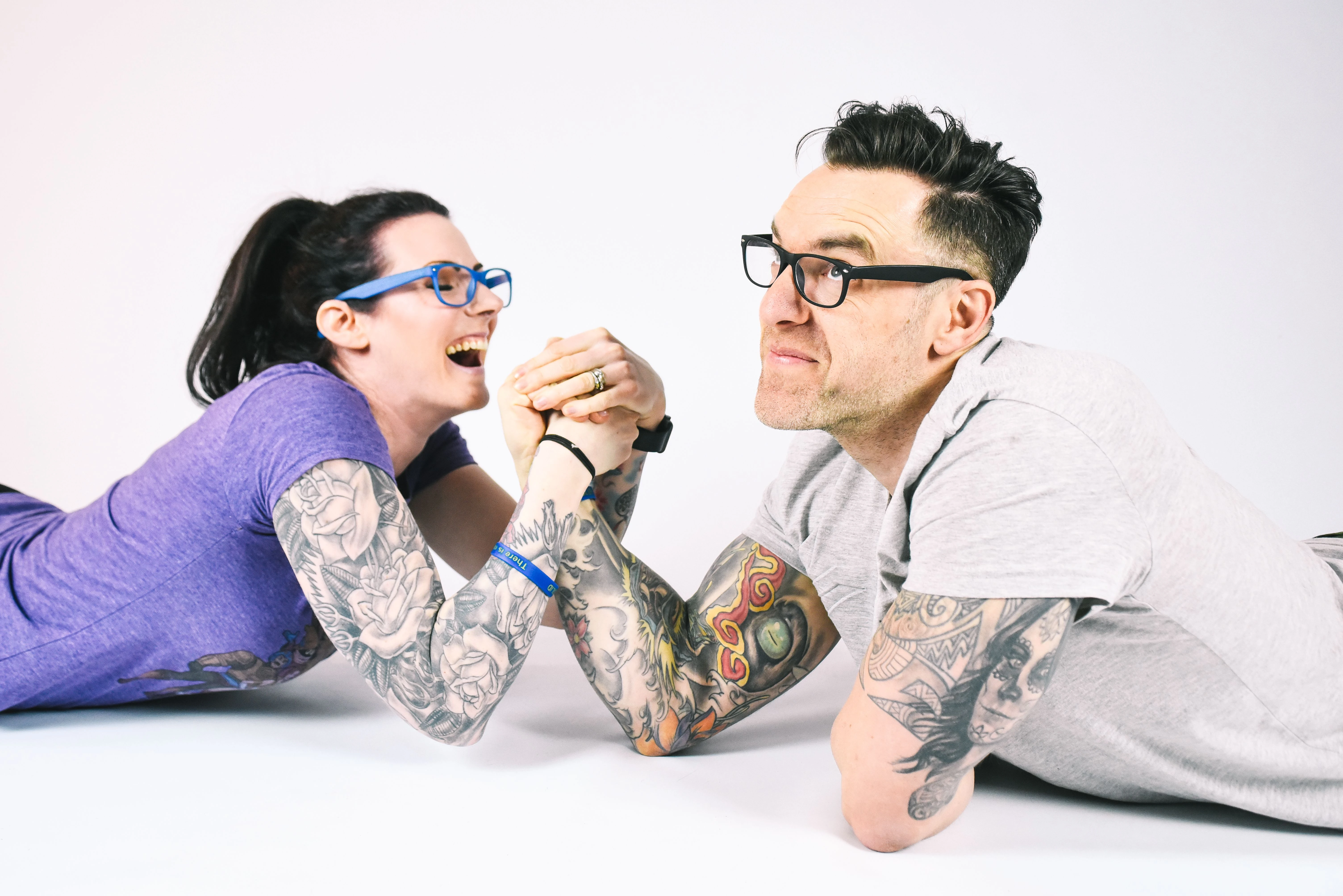 Loz and Baz arm-wrestling, laughing, against a plain studio backdrop