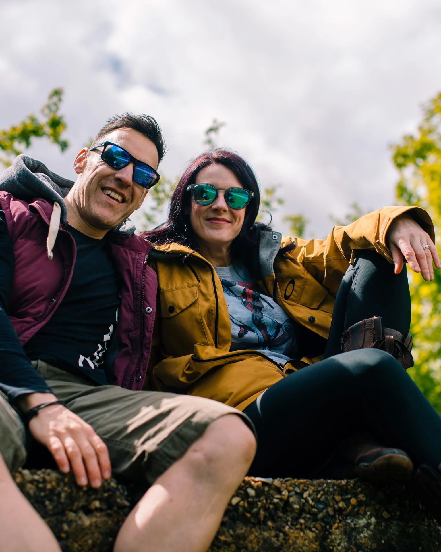 Loz and Baz sitting on a wall outdoors in sunglasses, laughing